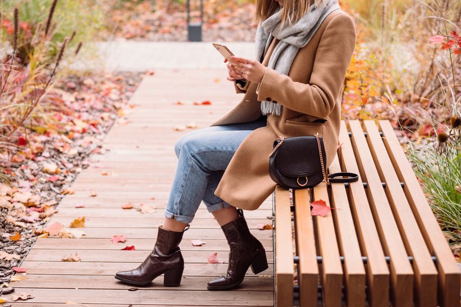 Elegant woman in a brown coat sits on a bench surrounded by autumn foliage, using her phone.