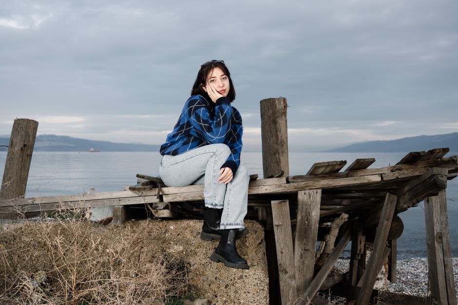 Woman in blue sweater and jeans sitting on a wooden structure by the sea. Overcast skies create a serene atmosphere.