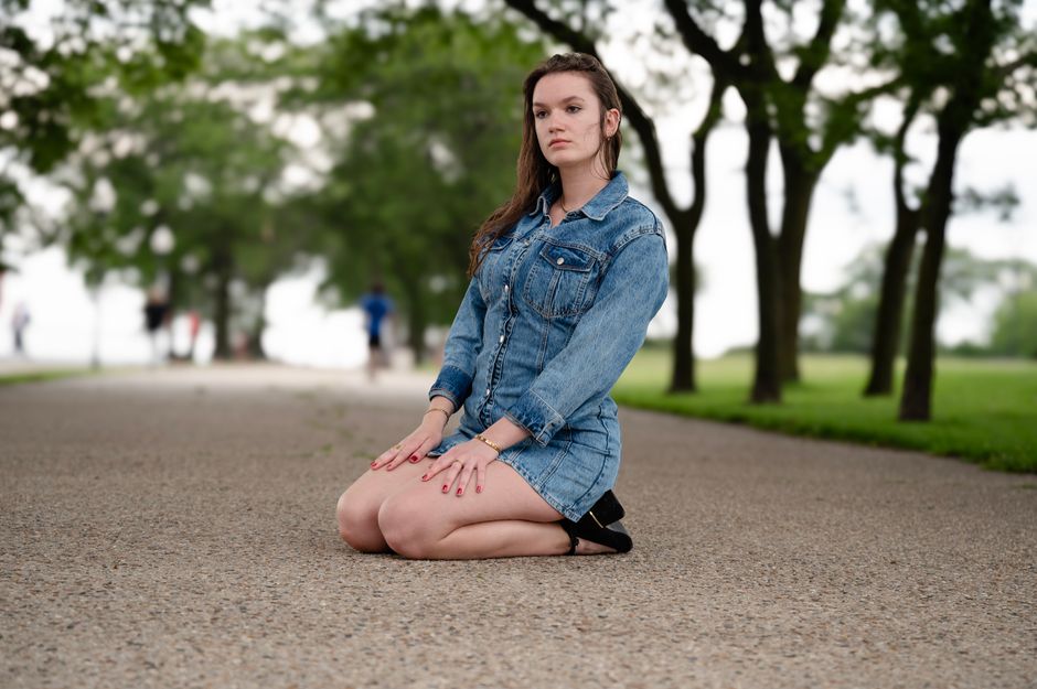 Young woman in denim kneels thoughtfully on a park pathway surrounded by greenery.