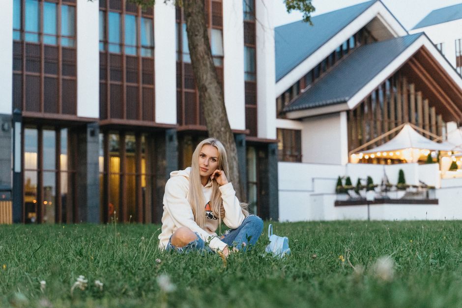 A young woman sitting on grass in front of a modern building with illuminated lights in Minsk, Belarus.