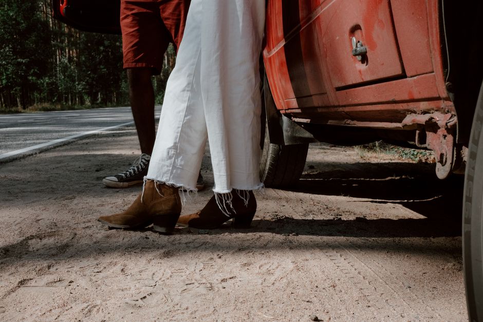 Close-up of casual footwear on a sandy roadside next to a red vehicle in daylight.