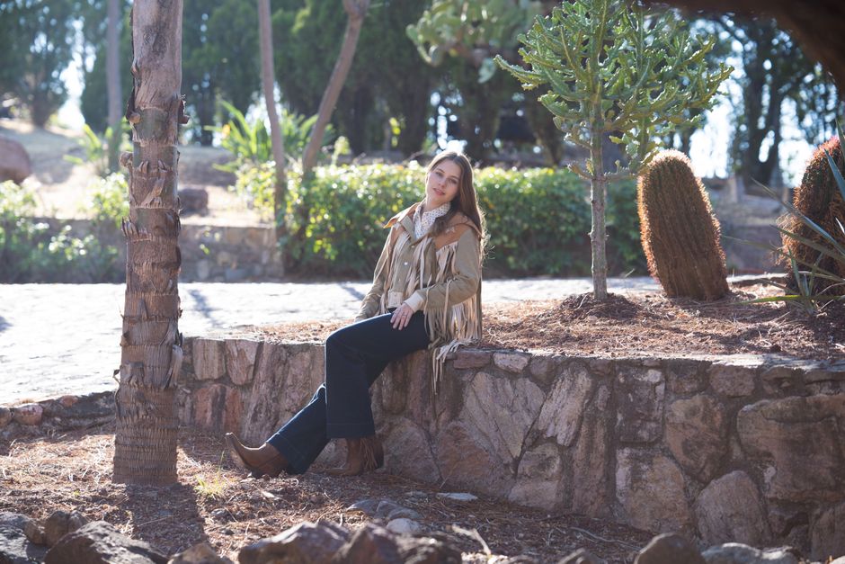 Woman in fringe jacket seated in sunny desert garden with cacti.