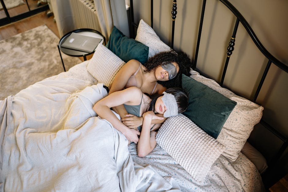 Two women in an affectionate embrace wearing sleep masks, enjoying a peaceful morning in bed.