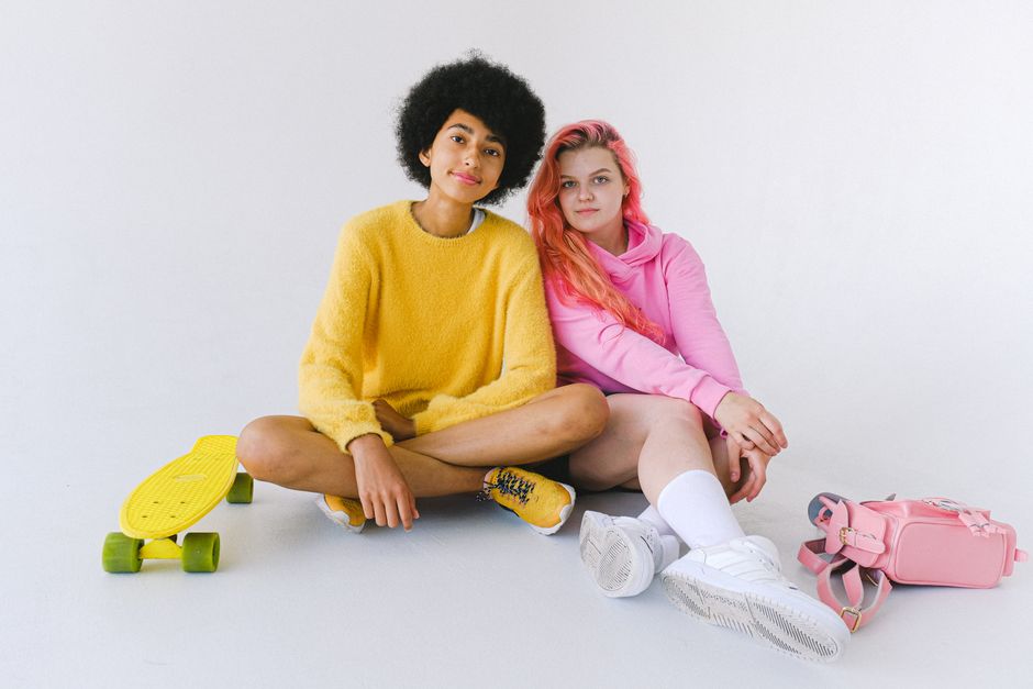 Full body of diverse friends in bright yellow sweater and pink sweatshirts sitting with penny board and backpack in white studio