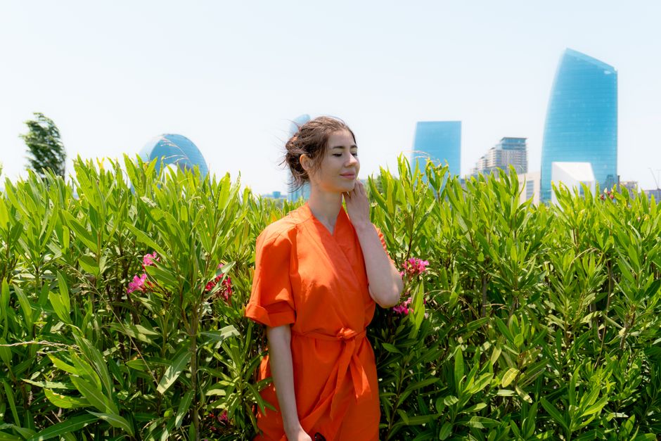 Woman in a vibrant orange dress stands amidst green foliage with modern city buildings in the background.