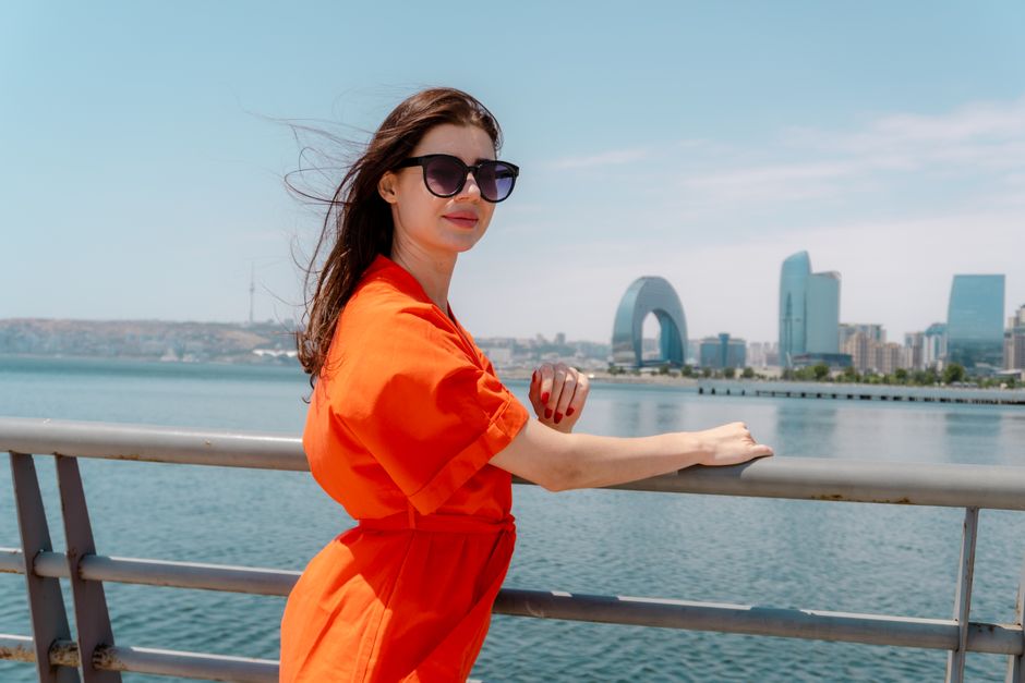 Woman in sunglasses and orange dress by waterfront with skyline view.