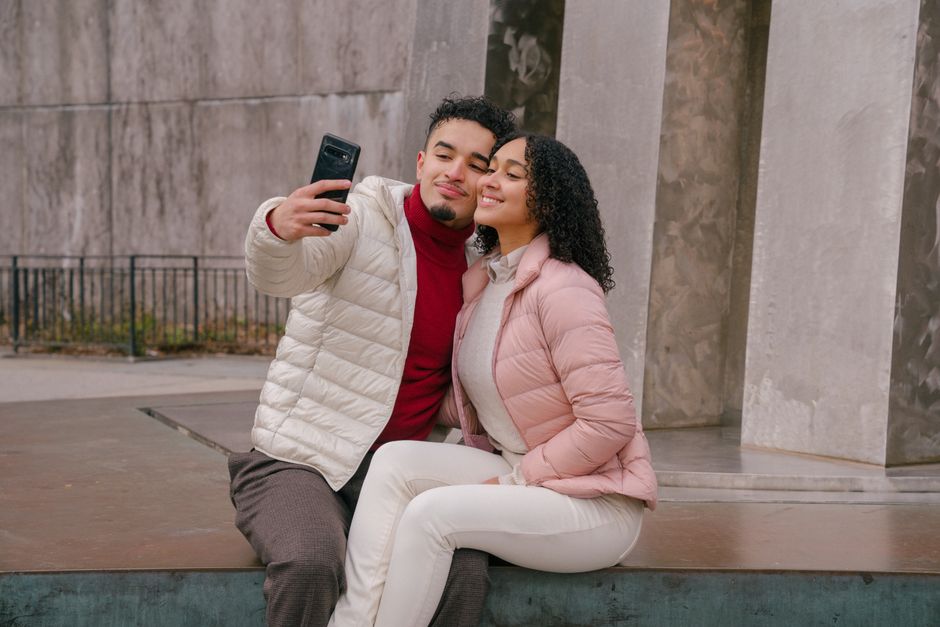 Content young Hispanic couple in cuddling and smiling while taking self portrait on smartphone sitting on stone parapet in autumn park