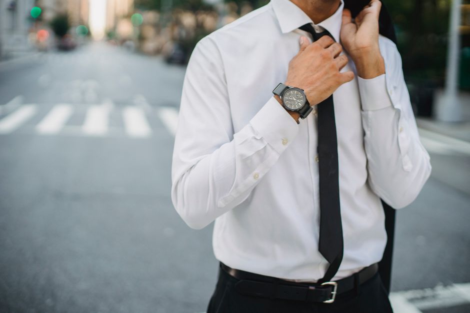 Confident businessman adjusting tie in a city street, wearing a classic black suit and watch.
