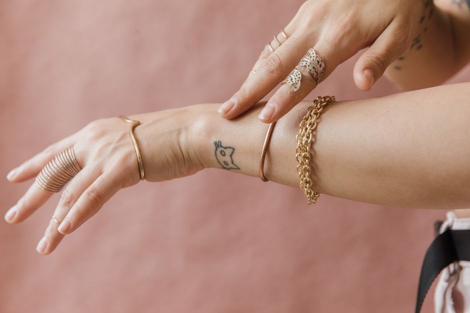 Close-up of a tattooed arm wearing elegant gold bracelets and rings on a soft background.