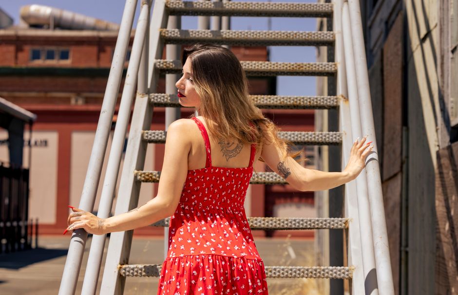 Stylish woman in a red sundress poses on an industrial staircase under the bright sun.