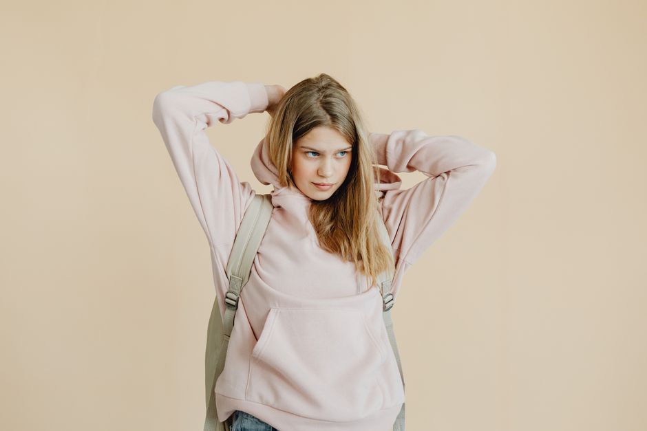 Teen girl wearing a pink hoodie and backpack, standing against a beige background.