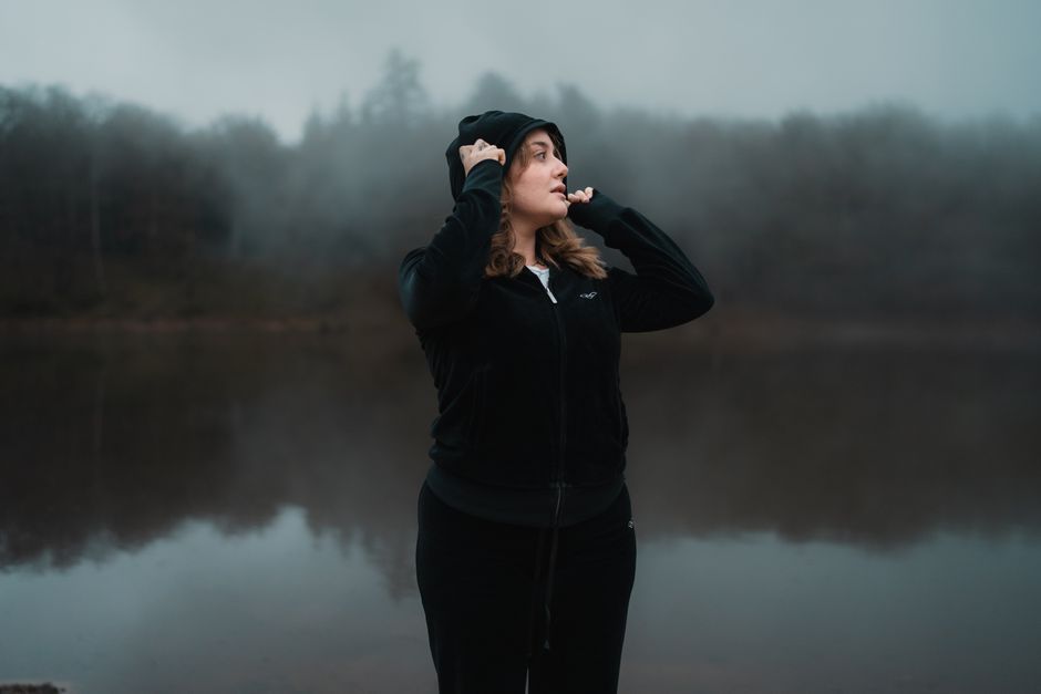 Woman in winter attire stands by a misty lake, exuding calm and introspection.