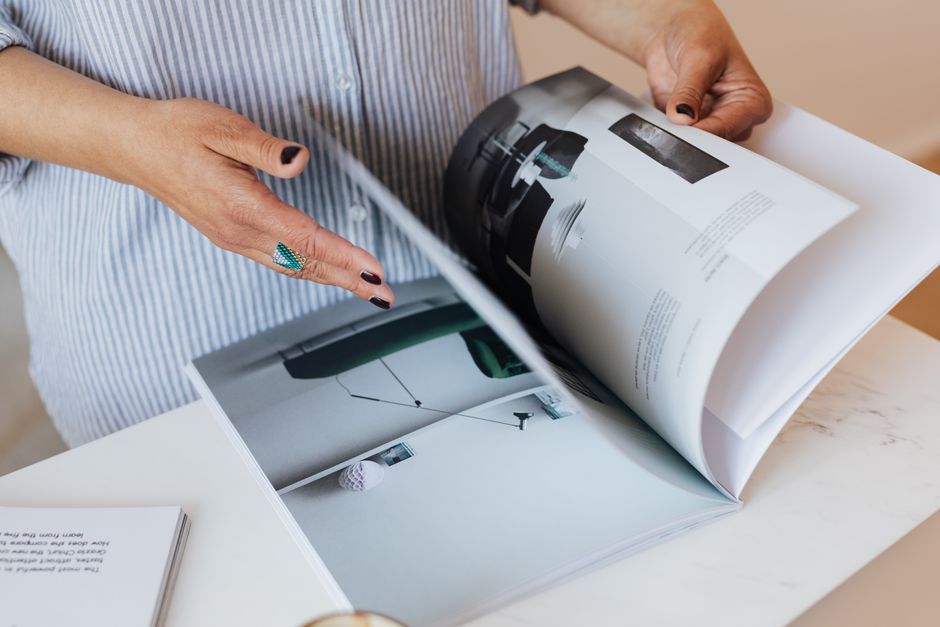 Crop unrecognizable female with manicured hands reading interesting magazine about interior design while standing near white marble table