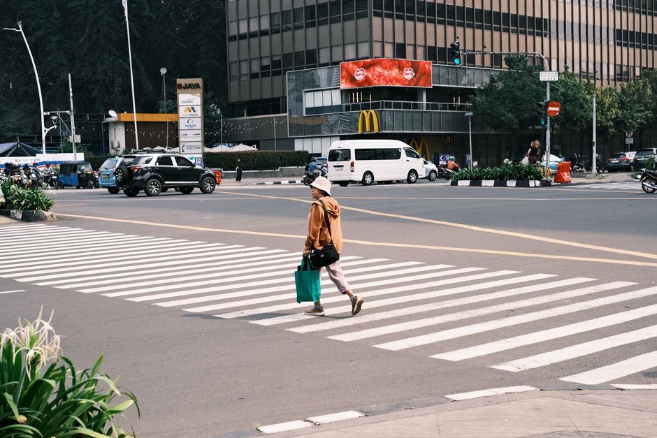 An adult walks across a city crosswalk near a McDonald's with modern buildings in the background.