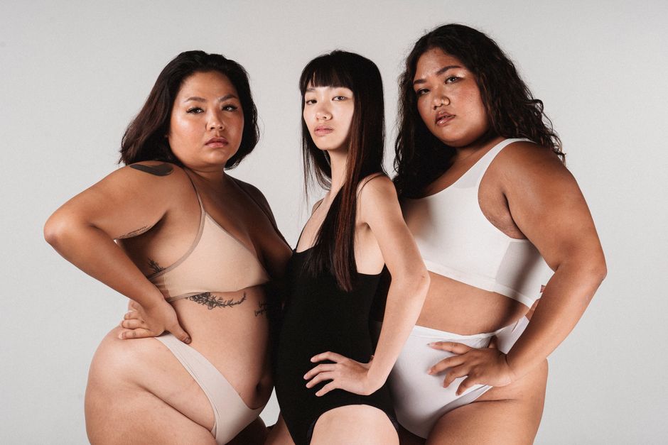 Asian slim woman in black bodysuit standing between plus size models in underwear and looking at camera while standing against white background with hand on waist