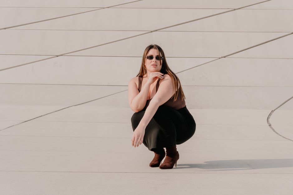 Stylish woman in sunglasses and boots poses against modern architectural backdrop.