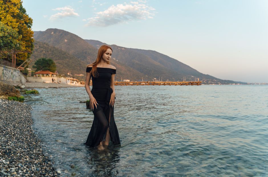 A woman in a black dress stands in the calm waters with mountain backdrop in Гагра.