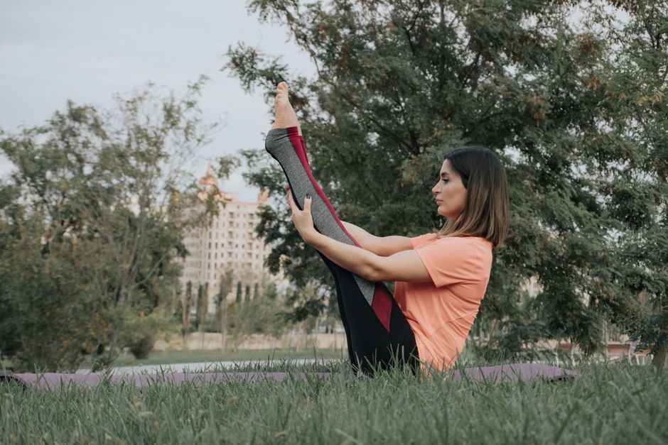 A woman performing a yoga stretch on grass in a serene park setting, promoting mindfulness and fitness.