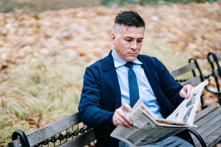 A man in formal attire absorbed in reading a newspaper while sitting on a park bench in fall setting.