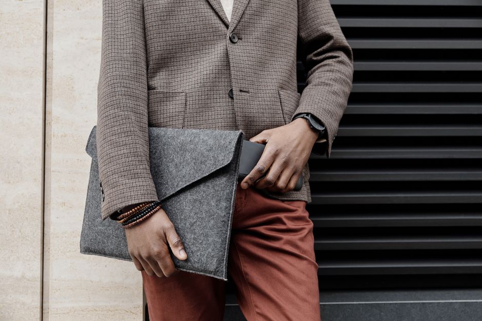 A stylishly dressed man in a brown jacket holds a gray felt bag, showcasing a modern fashion accessory.