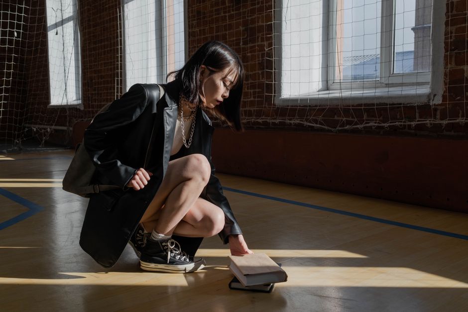Young woman in a black leather jacket squatting to pick up a book in an indoor sports hall.