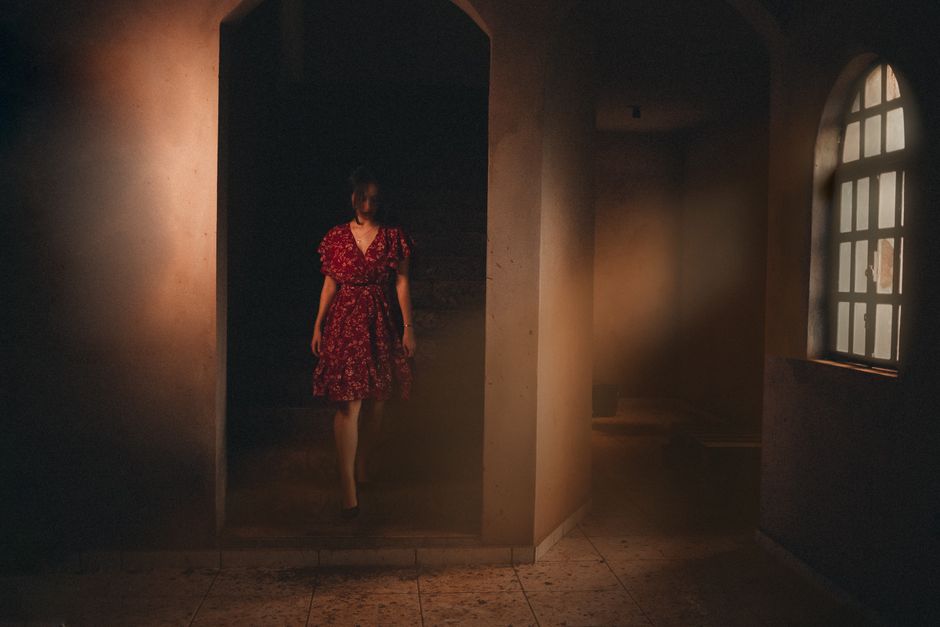 Atmospheric photo of a woman in a red dress standing in a dimly lit room.