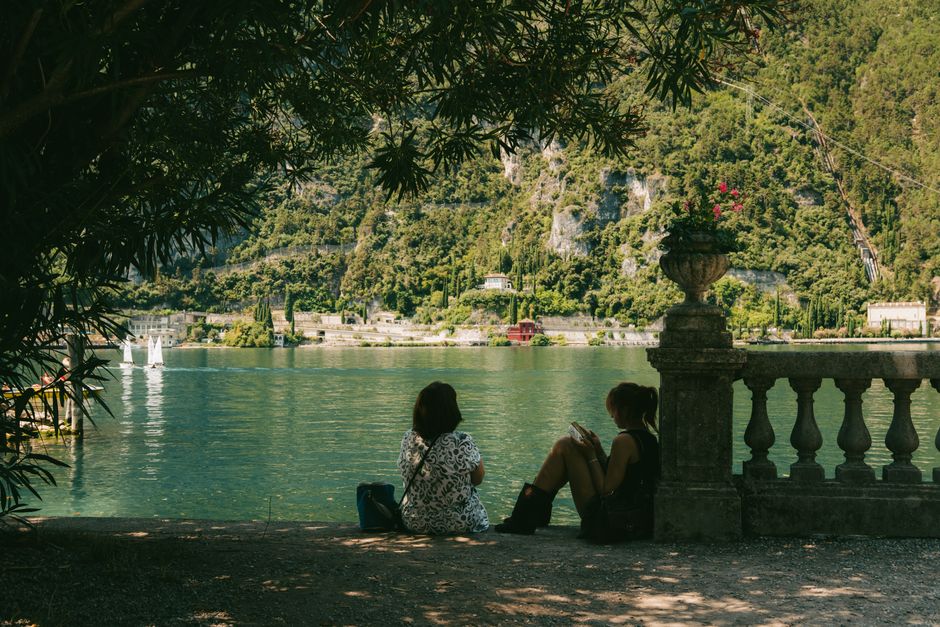 Two people relaxing by a serene Italian lakeside with sailing boats and lush greenery.