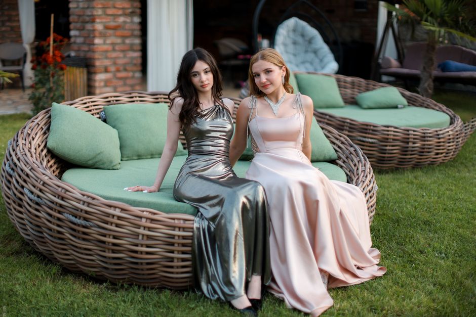 Two women in glamorous evening dresses relax on stylish outdoor furniture, exuding elegance and poise.