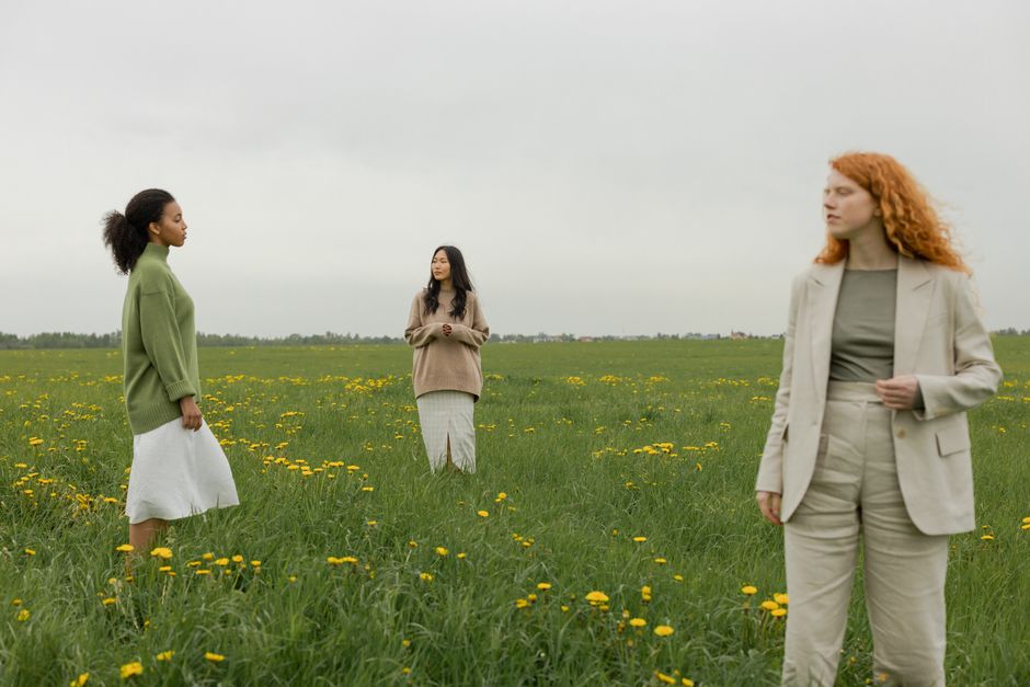 Three young women standing in a grassy field with yellow flowers under an overcast sky.