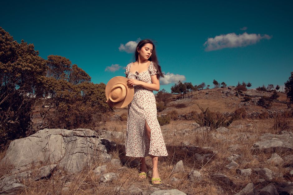 Young woman in a floral dress and hat standing in a rocky landscape.