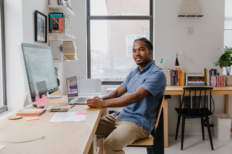 African American man sitting at a cluttered home office desk with computer and books.