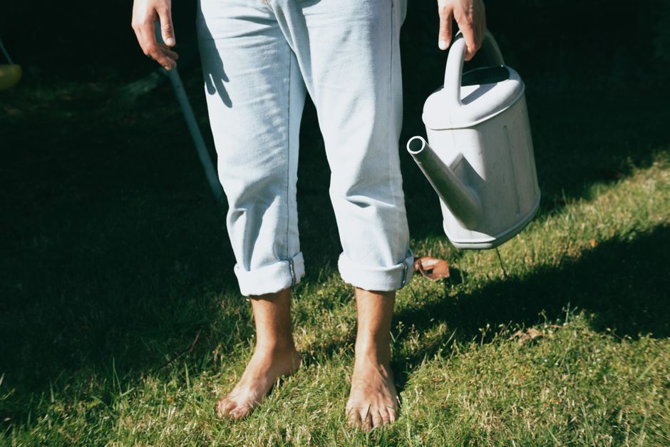 A person standing barefoot on grass holding a watering can, wearing rolled-up jeans in daylight.