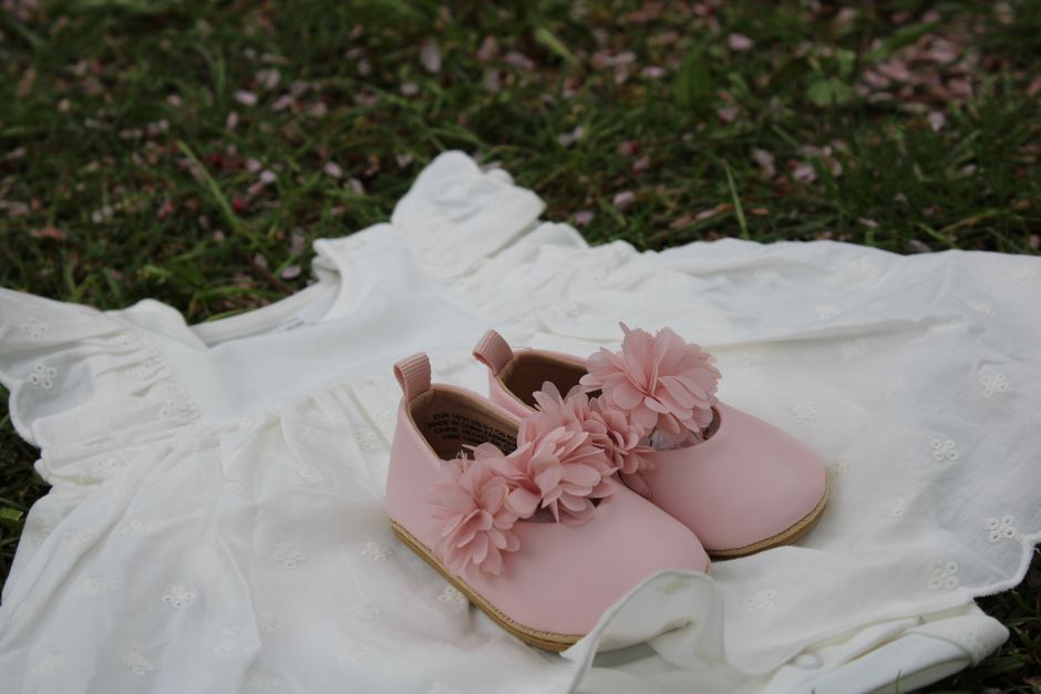 A delicate white baby dress paired with pink floral baby shoes on grass, perfect for spring time.