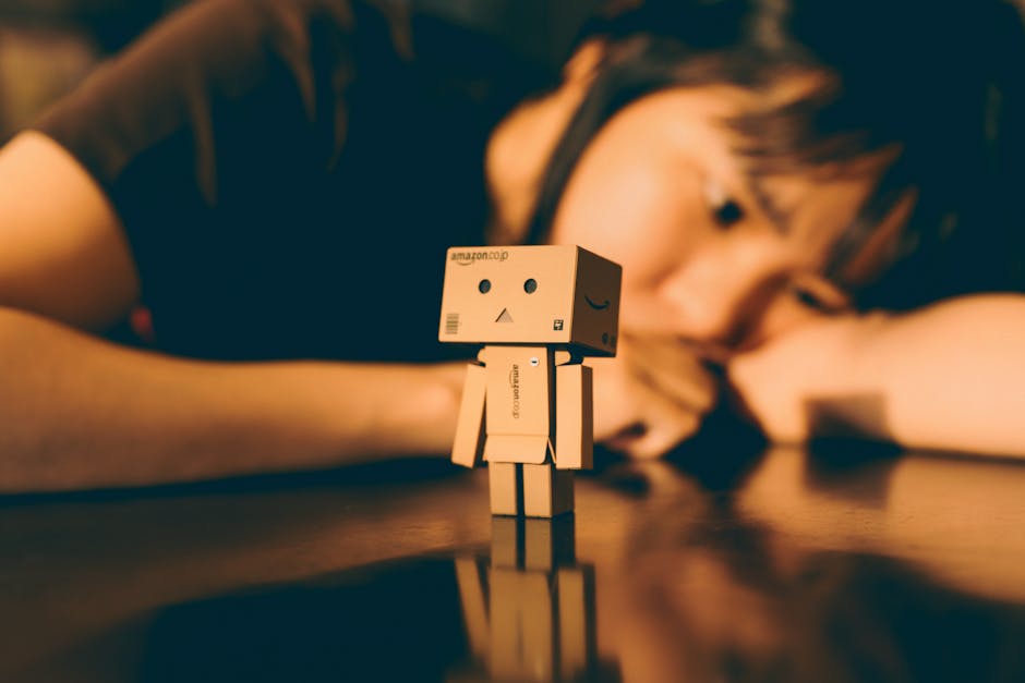 A Danbo cardboard robot stands on a reflective surface with a person in the background.