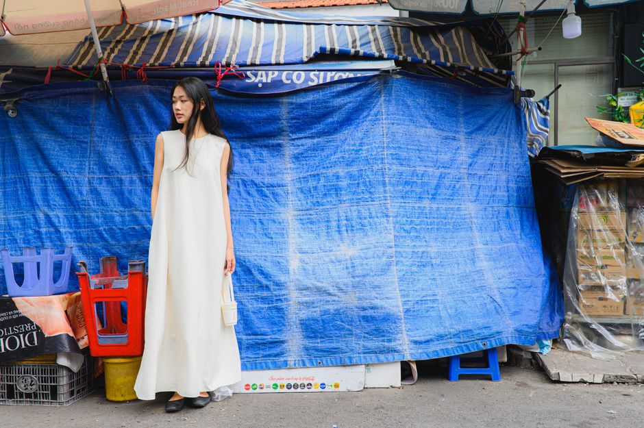 A fashionable woman in a white dress stands by a vibrant blue tarp market setup, embodying urban style.