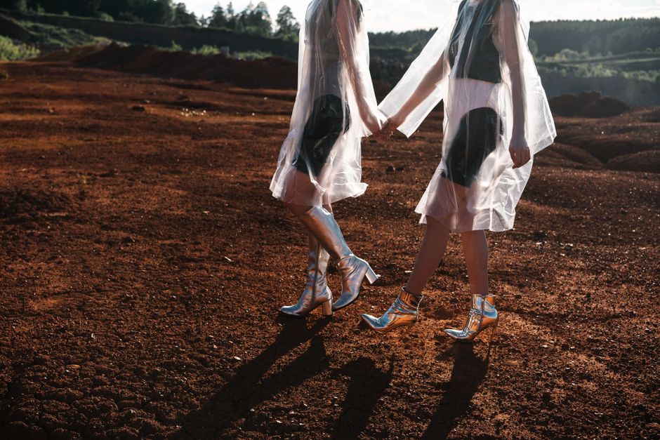 Two women in transparent outfits and silver boots walking in a red terrain setting.