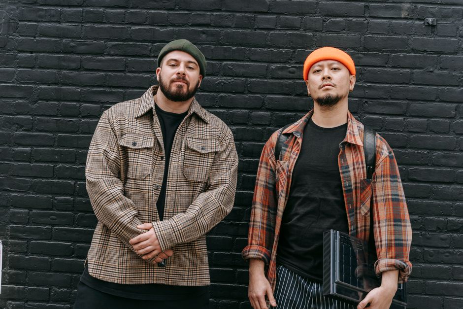 Two men in fashionable checkered jackets pose against a black brick wall outdoors.