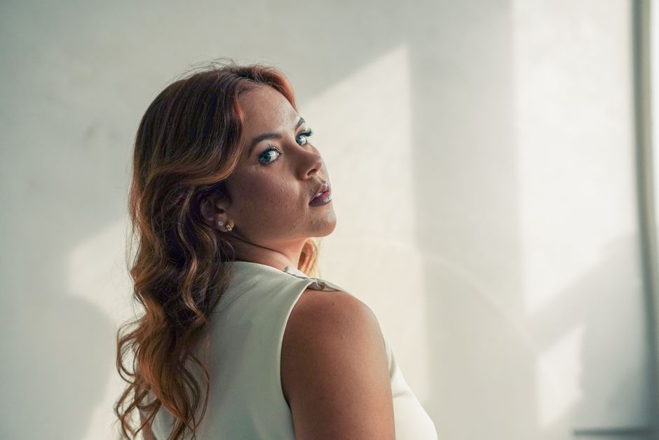 A woman with blue eyes looks over her shoulder, elegantly lit by soft sunlight indoors.