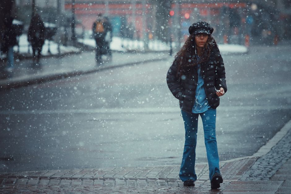 A woman walks through a snowy street in winter, wearing a jacket and hat.