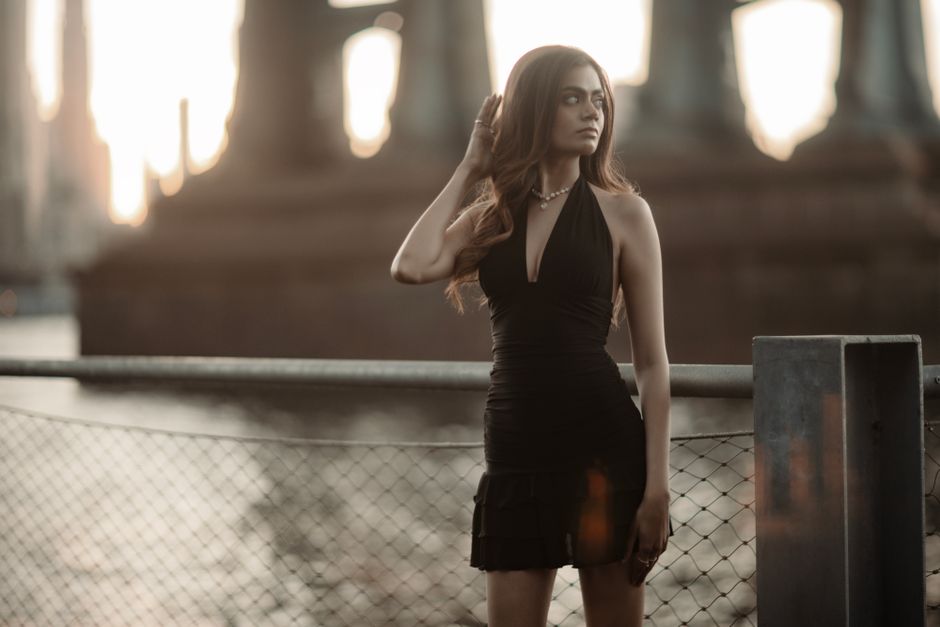 Woman in a black dress posing by a waterfront with a warm sunset backdrop, creating a serene mood.