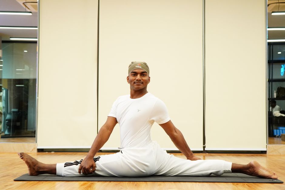 Athletic man in white sportswear doing splits on a mat in a gym setting, demonstrating flexibility.